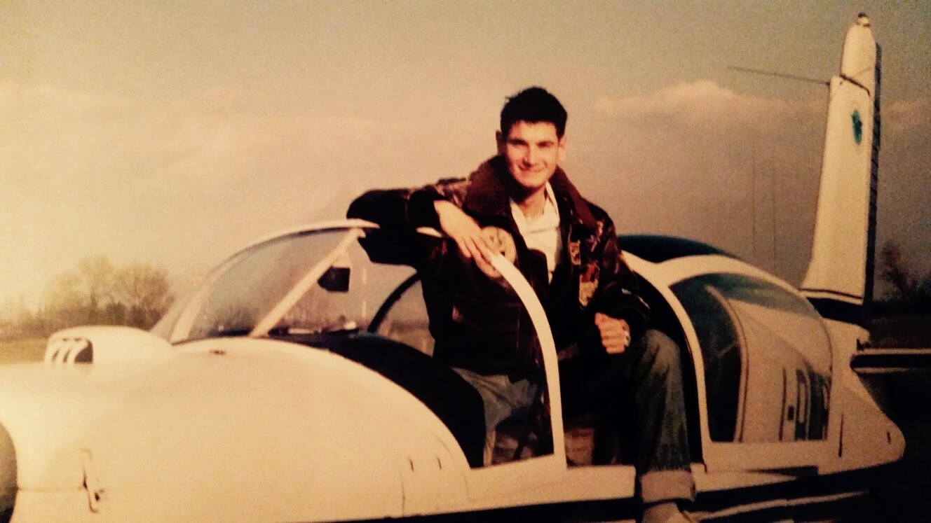 An early aviation photograph of a young man seated in an open light-aircraft cockpit