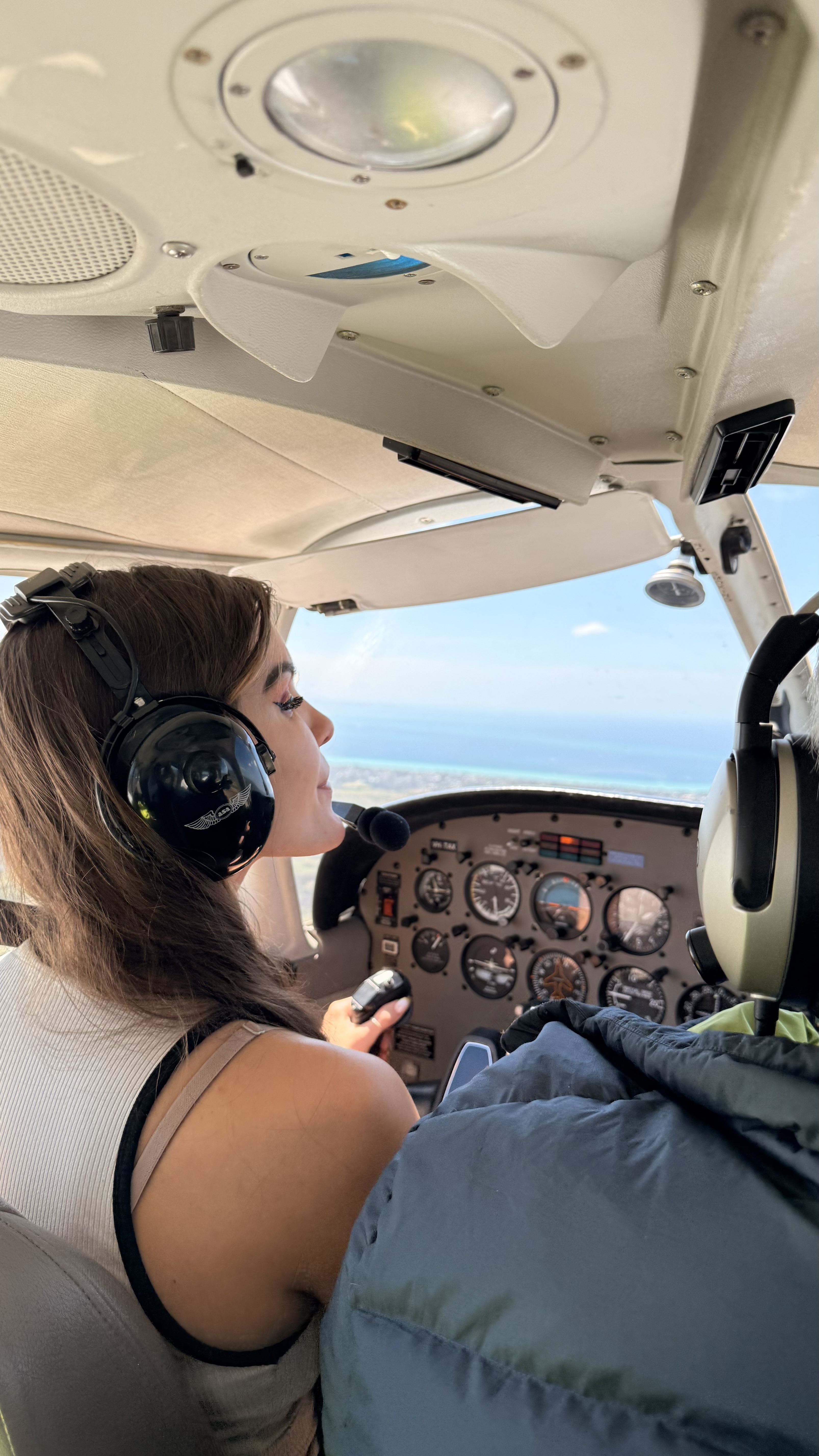 A headset-wearing person seated at a light-aircraft instrument panel above the coast