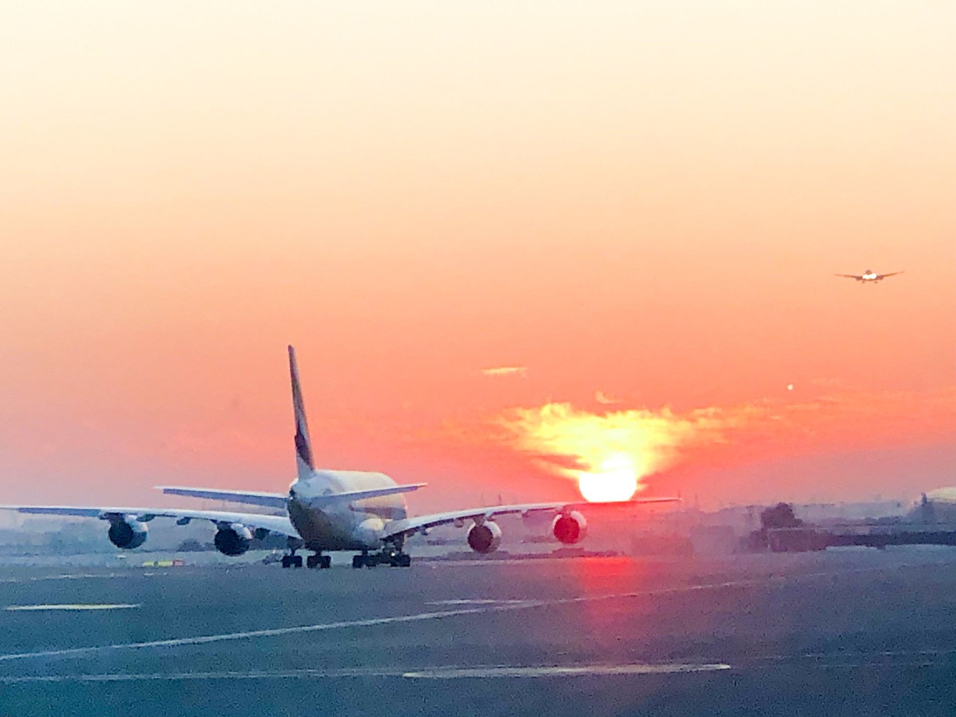 A jumbo jet taxiing towards the setting sun with another airliner above